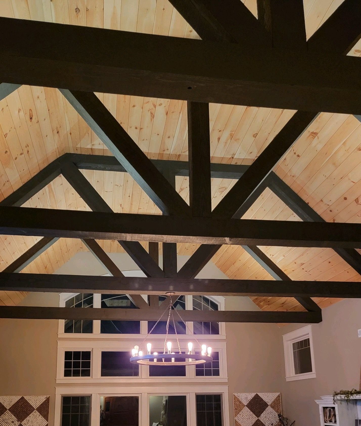 Wooden vaulted ceiling with dark beams, natural wood panels, and a chandelier below a wall of windows.