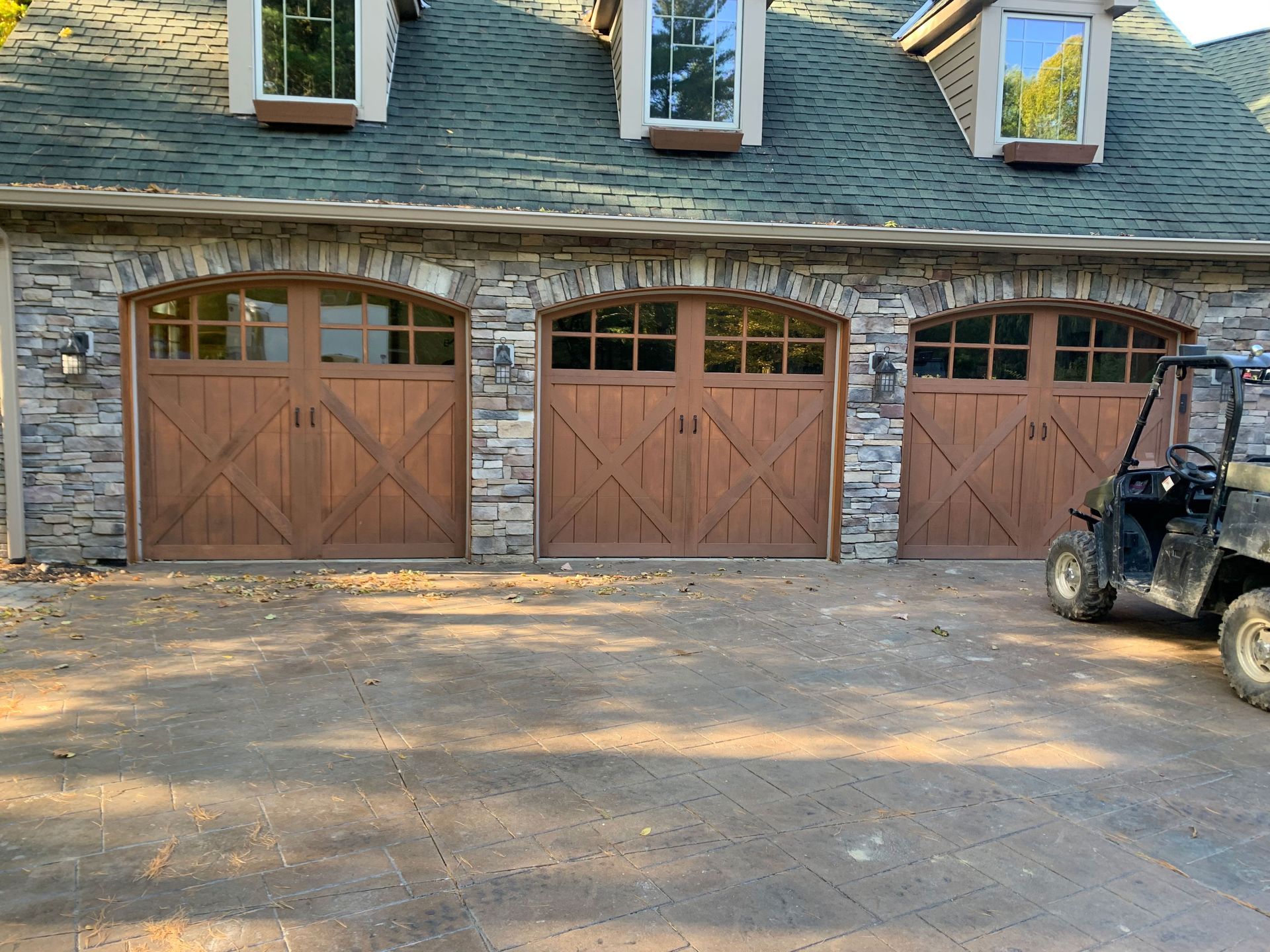 Three brown garage doors with stone archways and an ATV parked on a gravel driveway.