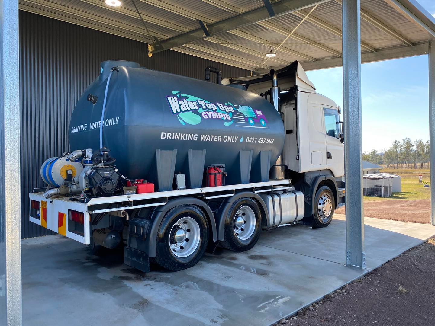 A gray septic truck under a gray metal roof, with a blue tank displaying business info, parked outside. — Water TopUps Gympie in Pie Tamaree, QLD