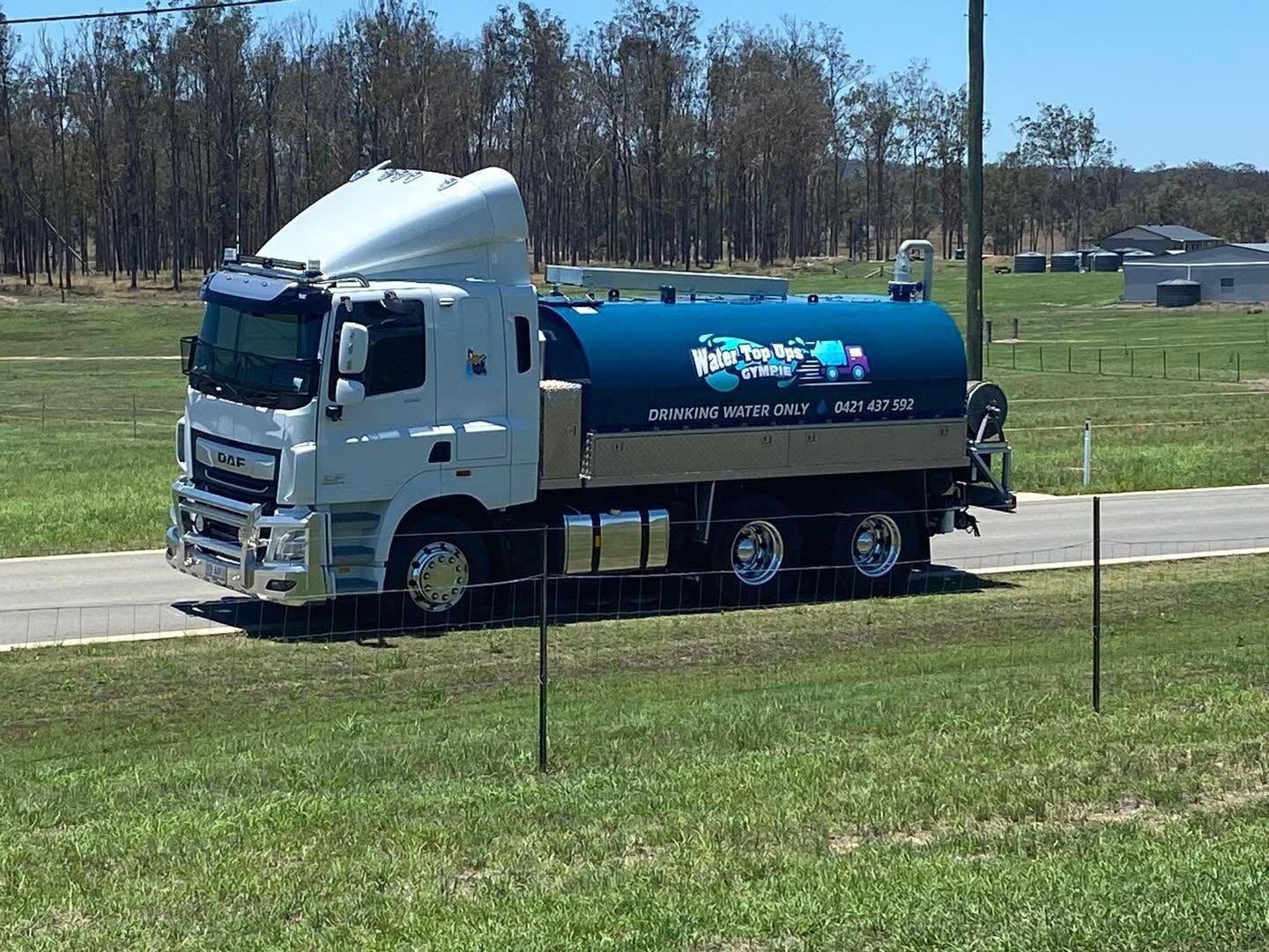White water tanker truck driving on a grassy roadside with trees in the background