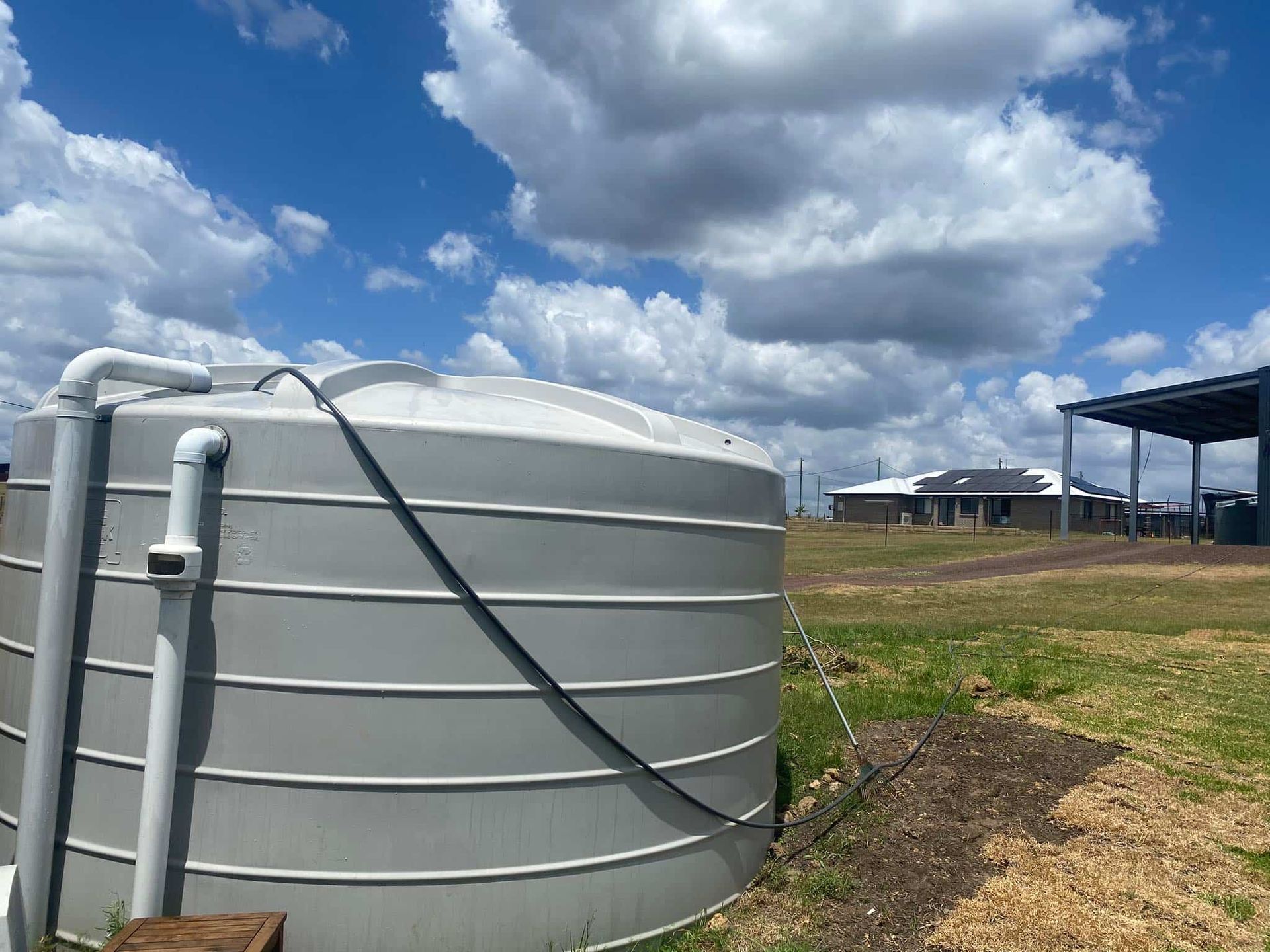 Large, grey water tank in grassy field with blue sky and some houses in the distance. — Water TopUps Gympie in Araluen, QLD