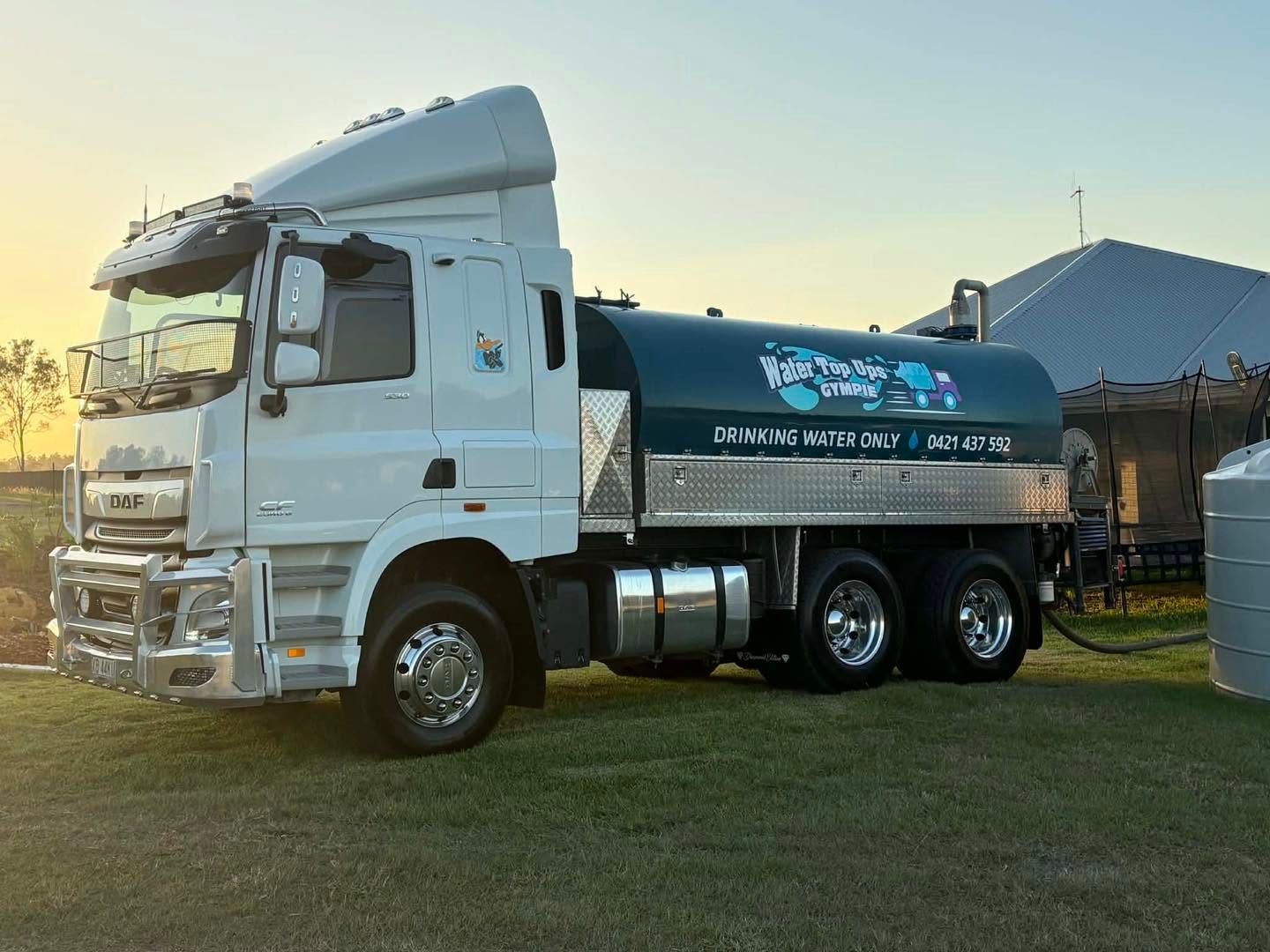 White tanker truck parked on grass at sunset beside a barn and silo