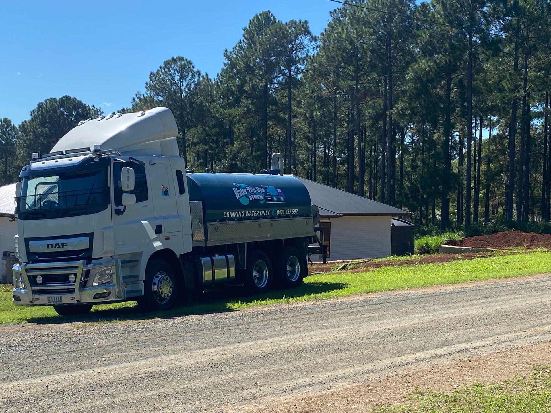 White tanker truck parked on a gravel road beside a small building and pine trees in bright daylight