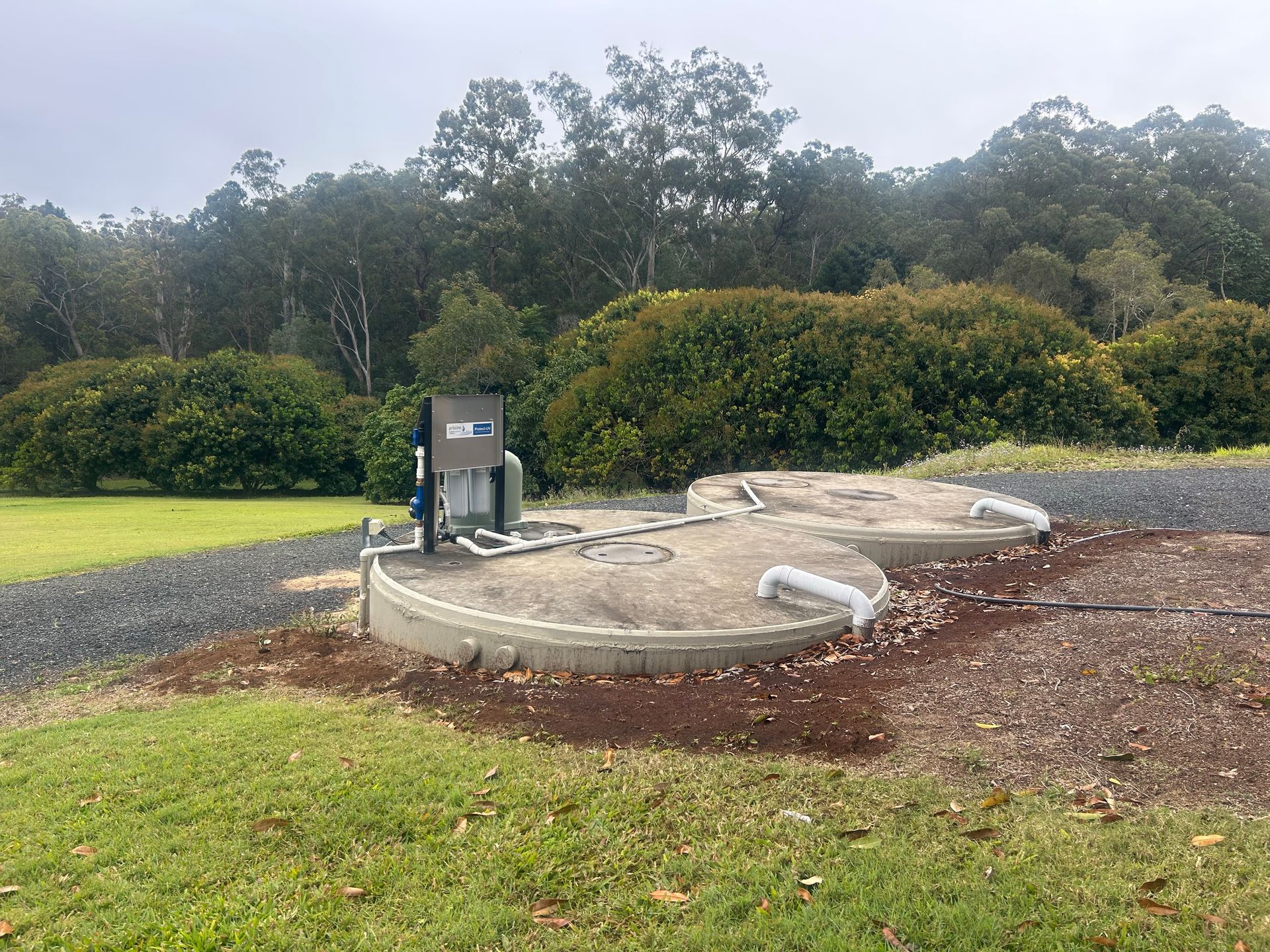 Outdoor utility equipment on a concrete pad beside a grassy area and trees, with pipes and a control box.