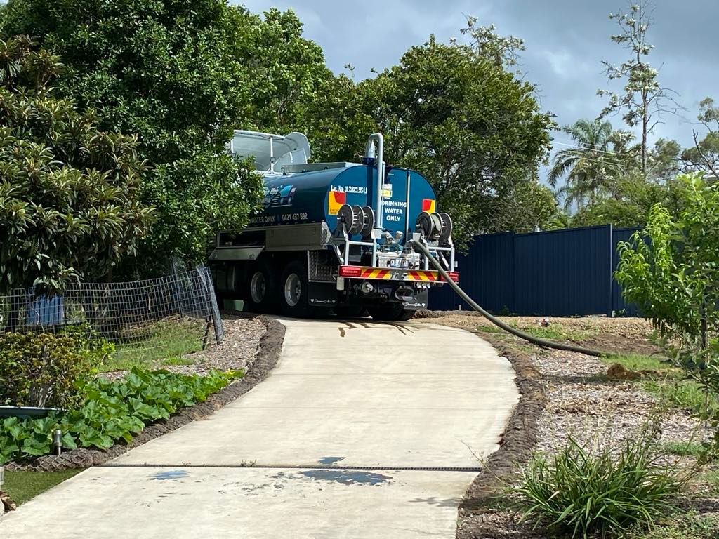 A blue septic tank truck parked on a concrete driveway, hose extending towards a blue fence, surrounded by green trees and foliage. — Water TopUps Gympie in Curra, QLD