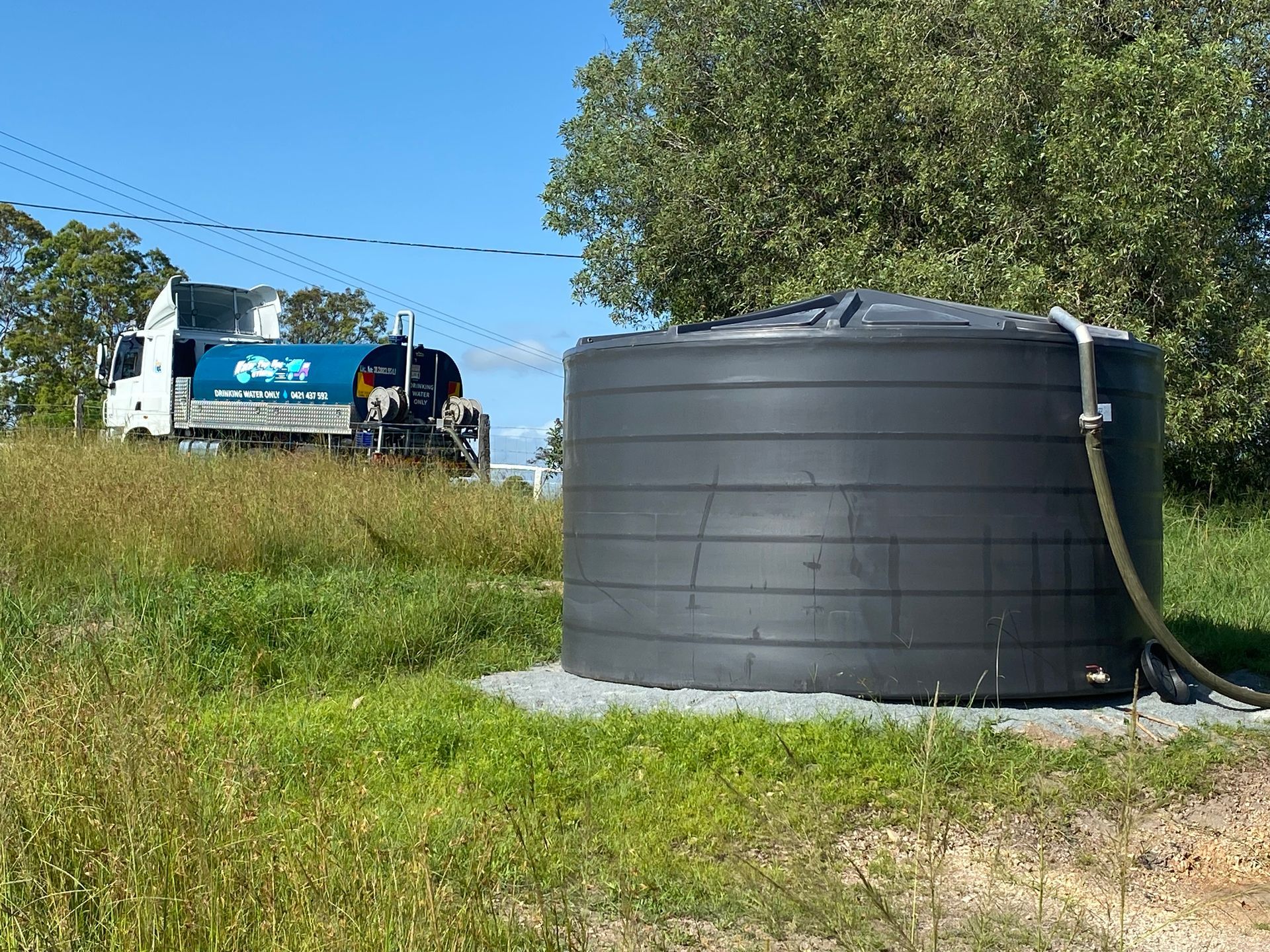 Large Black Water Tank in A Grassy Field with A Blue Tanker Truck — Water Top Ups Gympie in Nahrunda, QLD