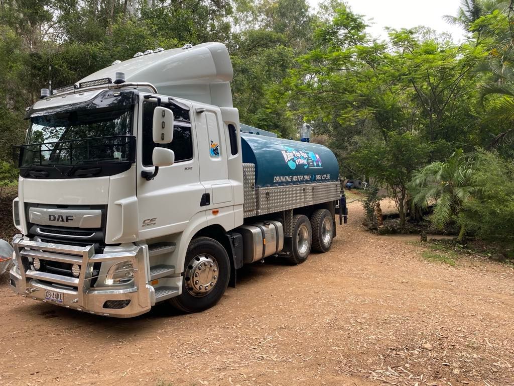 White tanker truck parked on a dirt road beside green trees and brush