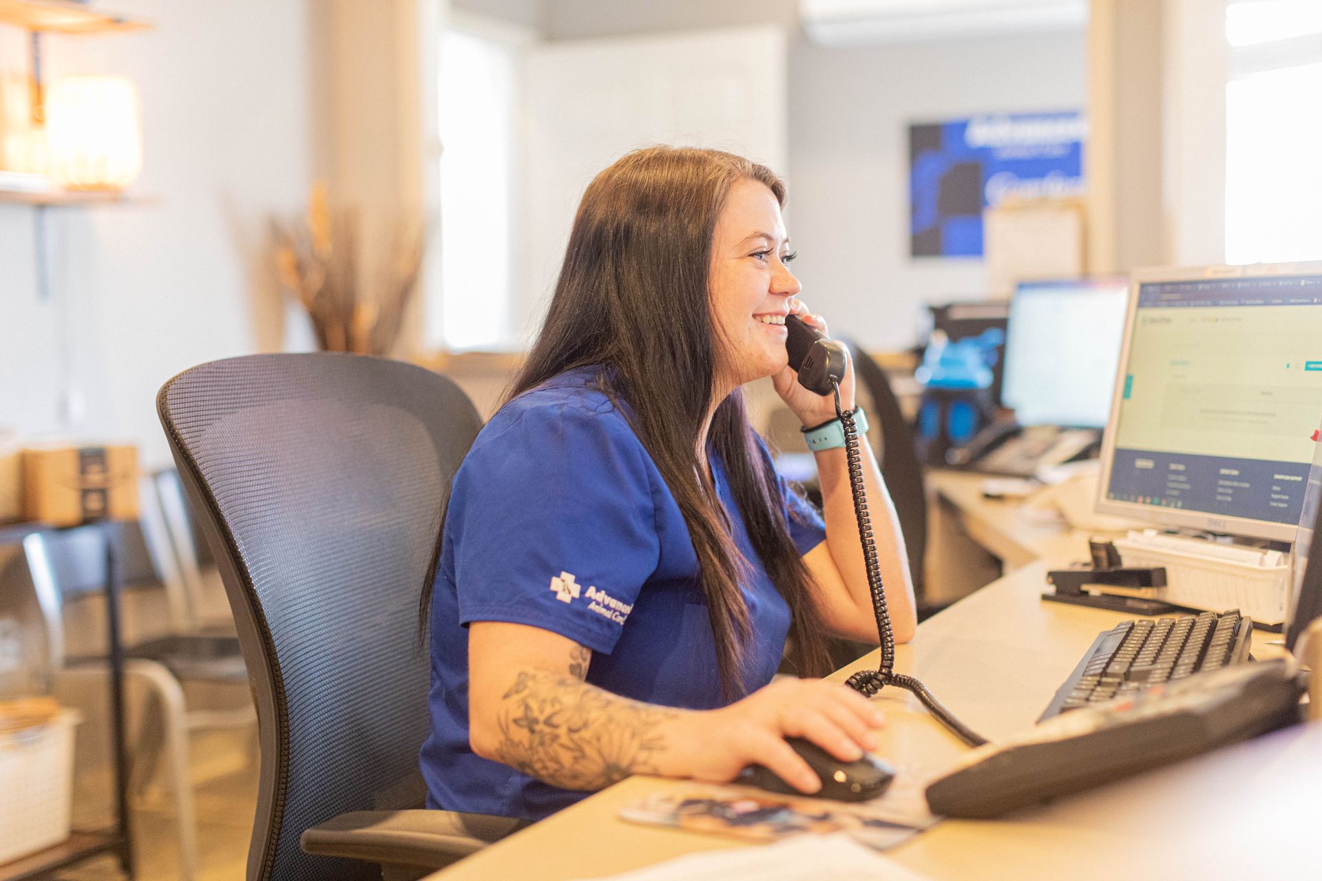 Woman in blue scrubs smiles while on the phone, working at a desk with computer and keyboard.