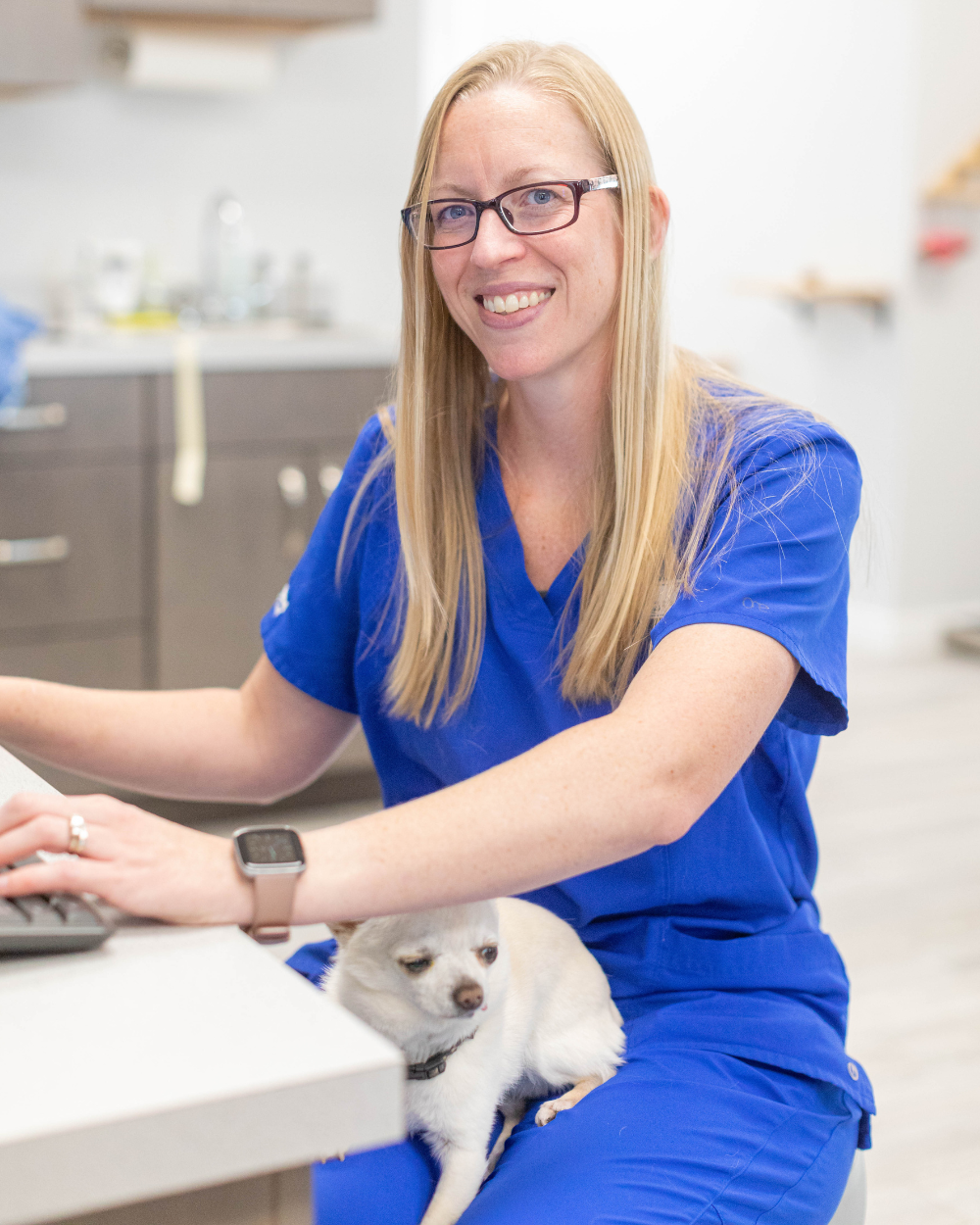 Woman in blue scrubs smiles while typing, a small white dog sits on her lap in an office.