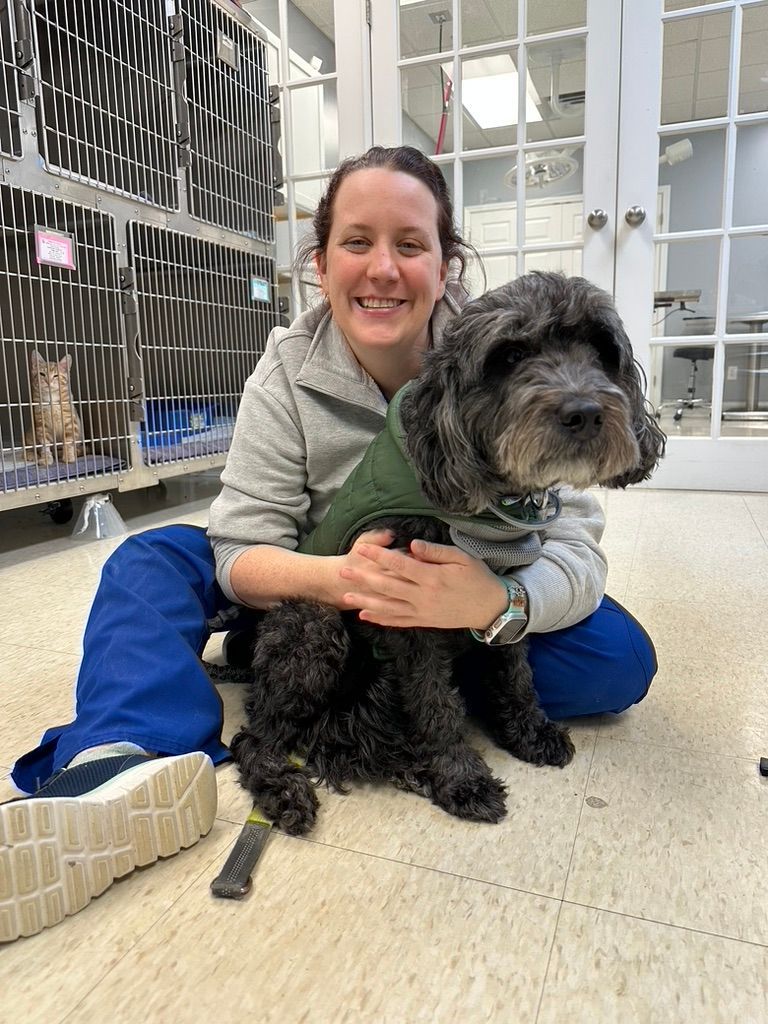Woman with curly hair smiles, sits next to brown and white dog in front of a brick wall.