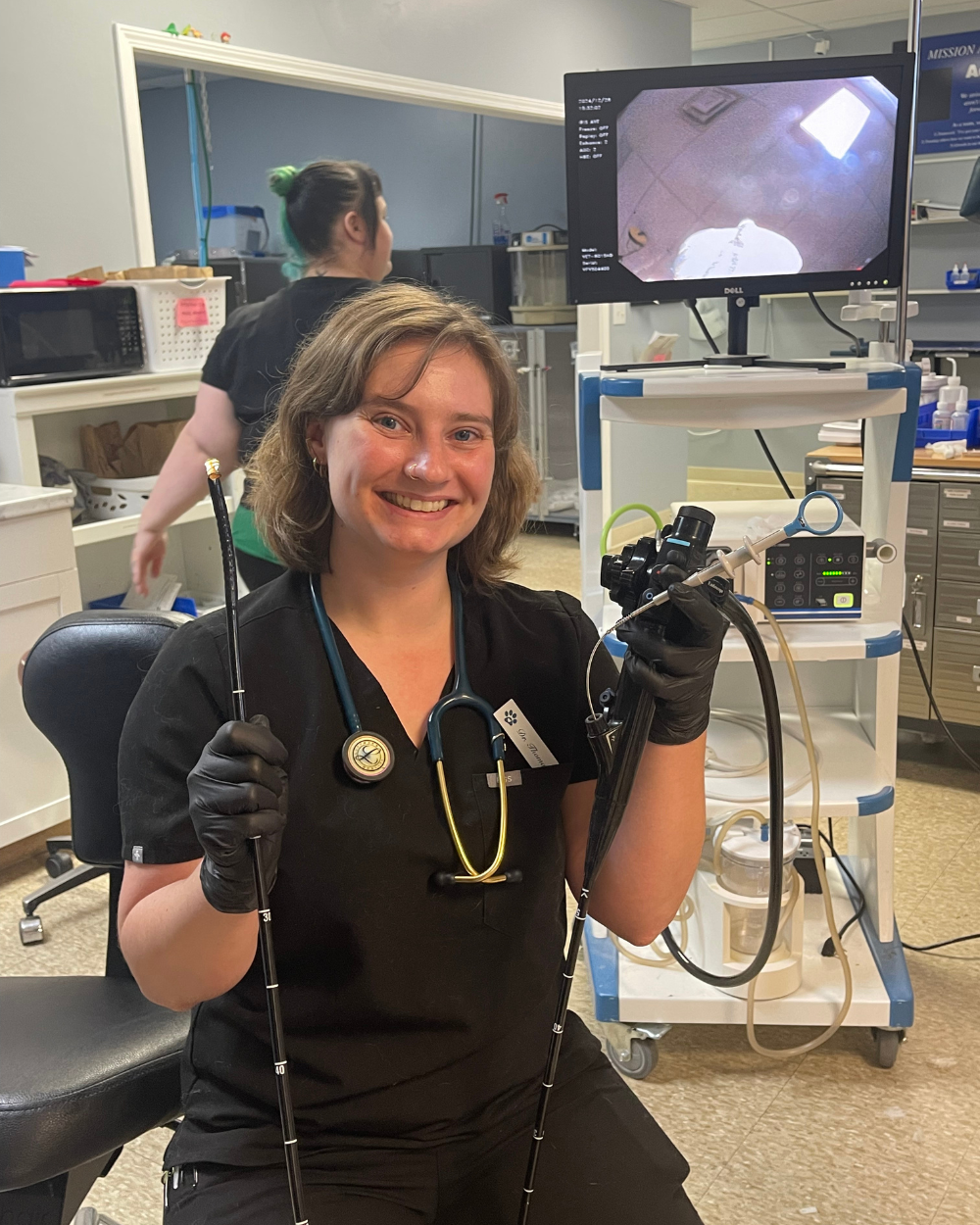 Veterinarian smiling, holding an endoscope. Monitors a stomach scope on a screen. Wearing black scrubs, gloves.