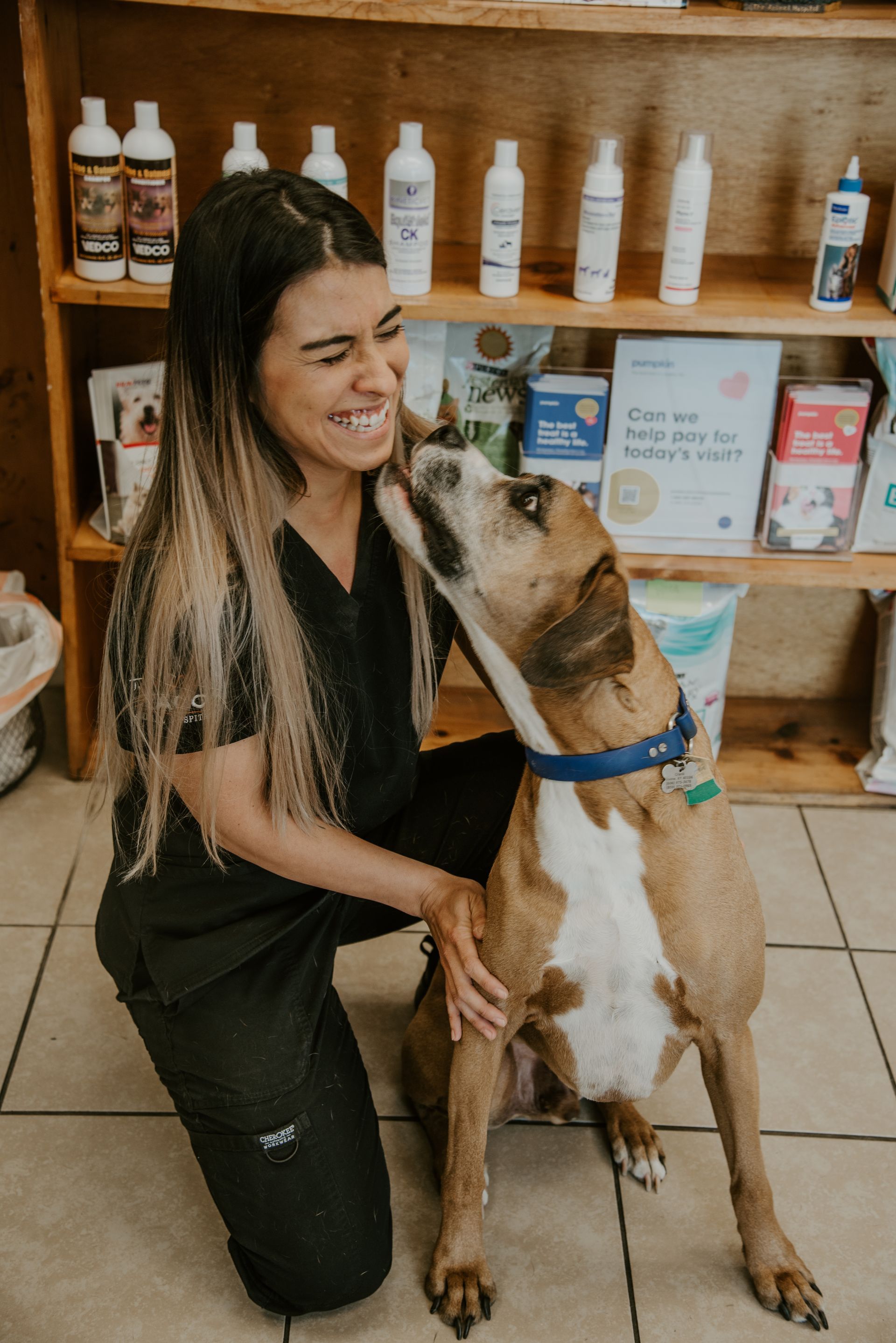 A woman in scrubs smiles as a dog licks her face in a pet supply store.