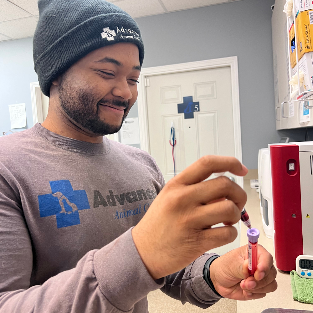 Man in gray shirt and beanie smiling, holding blood vials. Medical setting.