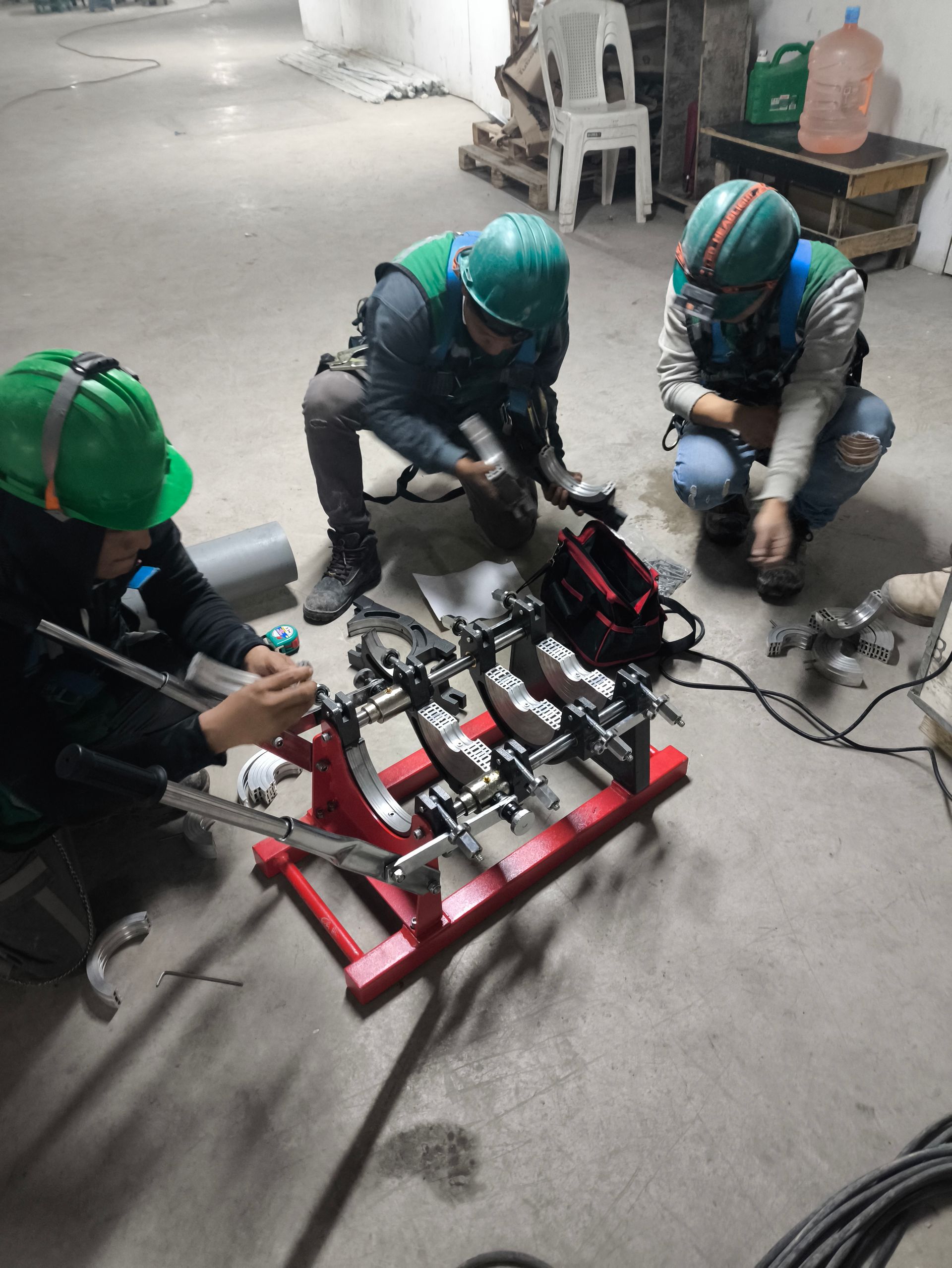 Trabajadores con cascos verdes ensamblan maquinaria sobre un suelo de hormigón con herramientas y cables.