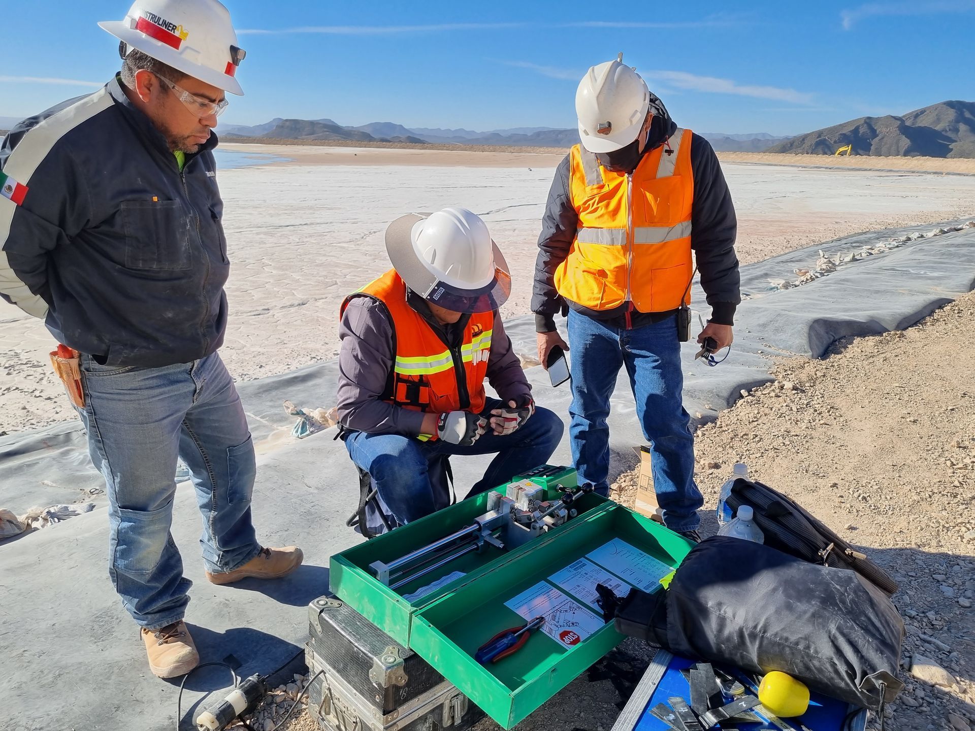 Obreros con cascos inspeccionan equipos junto a una salina y montañas.