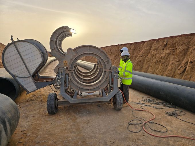 Obrero con chaleco reflectante junto a una gran máquina de tuberías metálicas en una obra de construcción al atardecer.