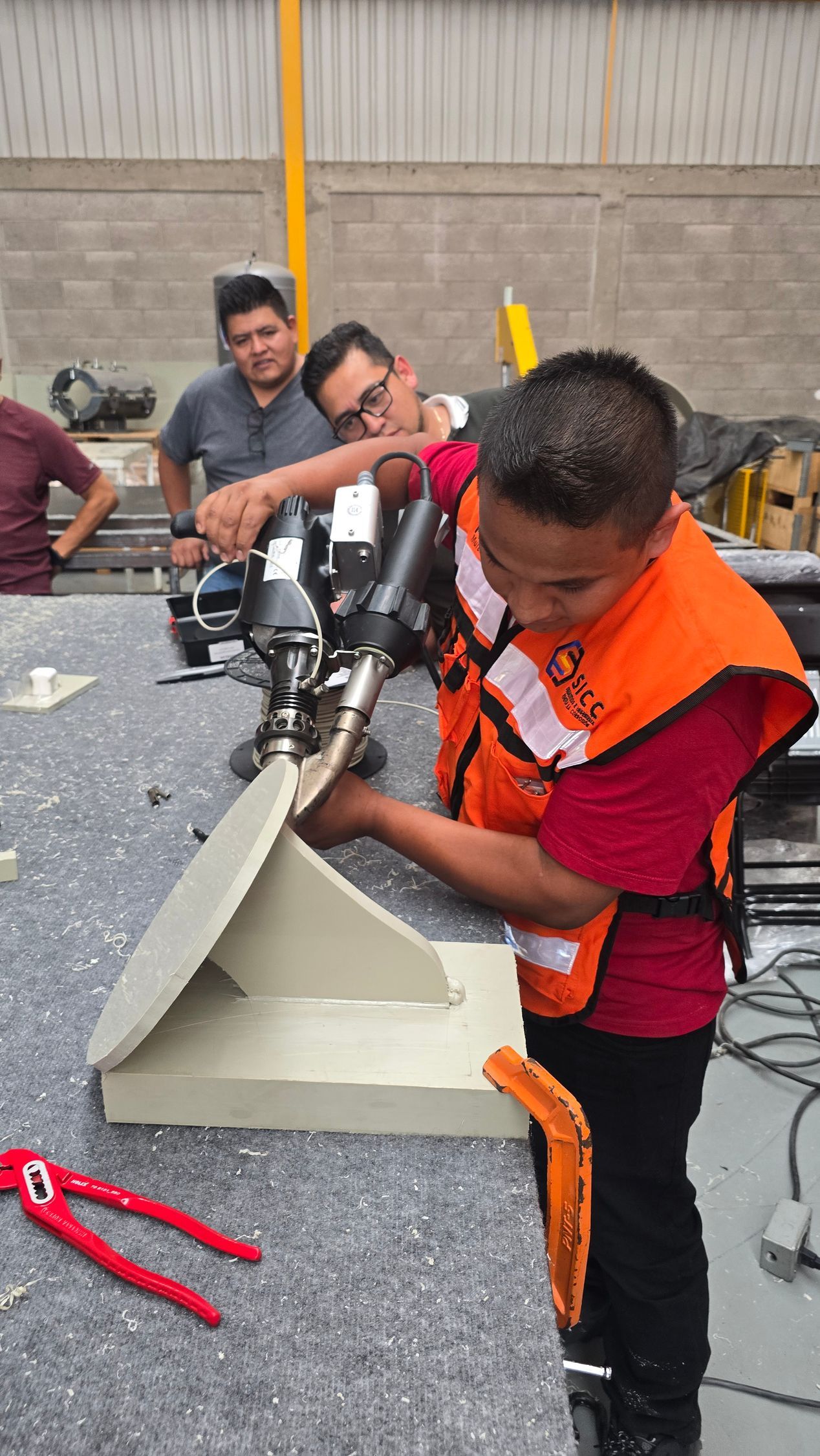 Varias personas trabajan con herramientas eléctricas sobre una lámina de metal en un taller; una de ellas, con un chaleco naranja, está inclinada sobre la pieza de trabajo.