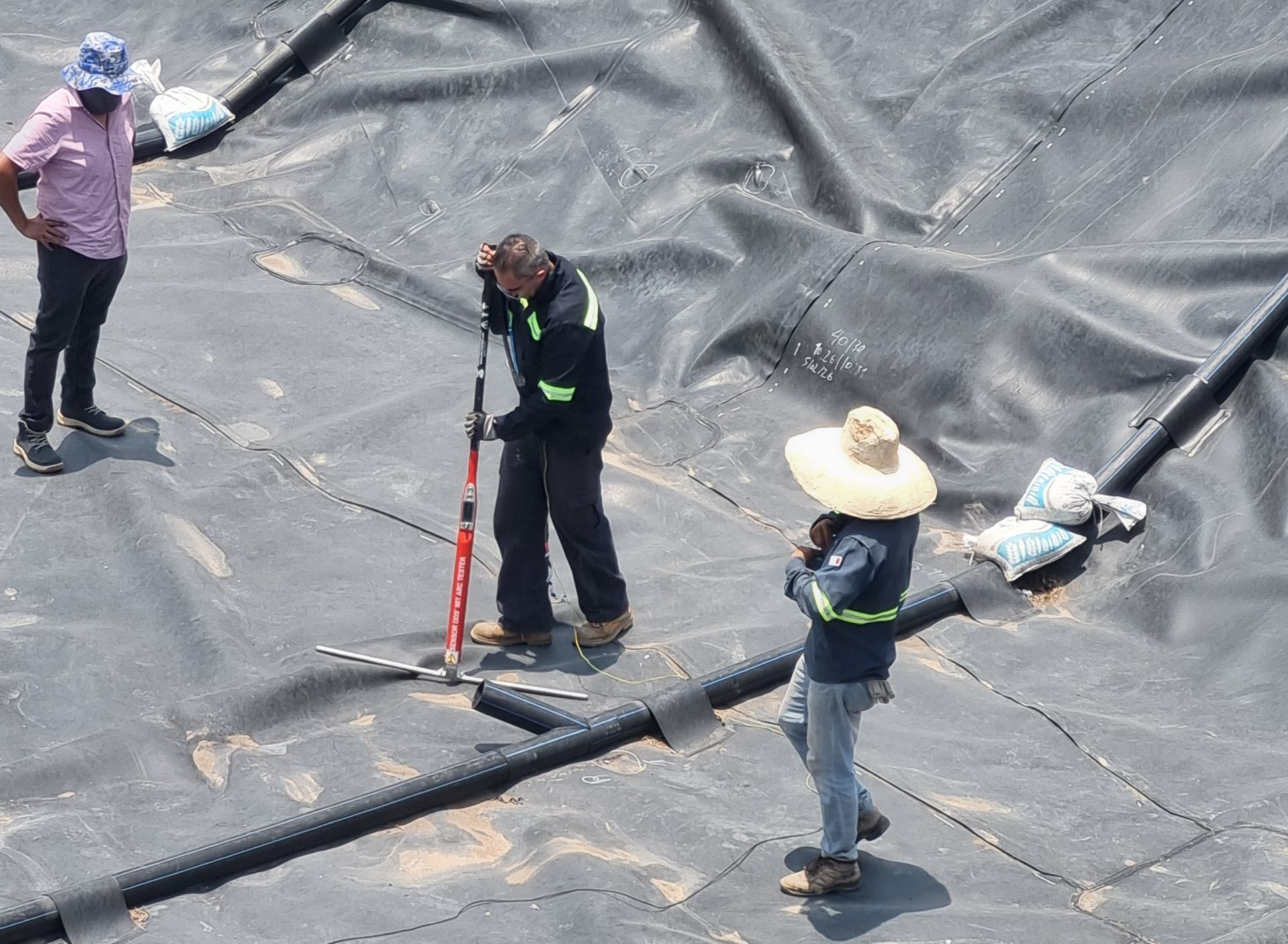 Tres trabajadores subidos a un tejado cubierto con una lona negra, utilizando herramientas y manipulando materiales cerca de la cumbrera.