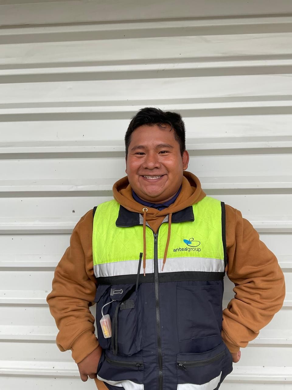 Persona sonriente con un chaleco de trabajo amarillo neón y azul marino de pie contra una pared blanca.