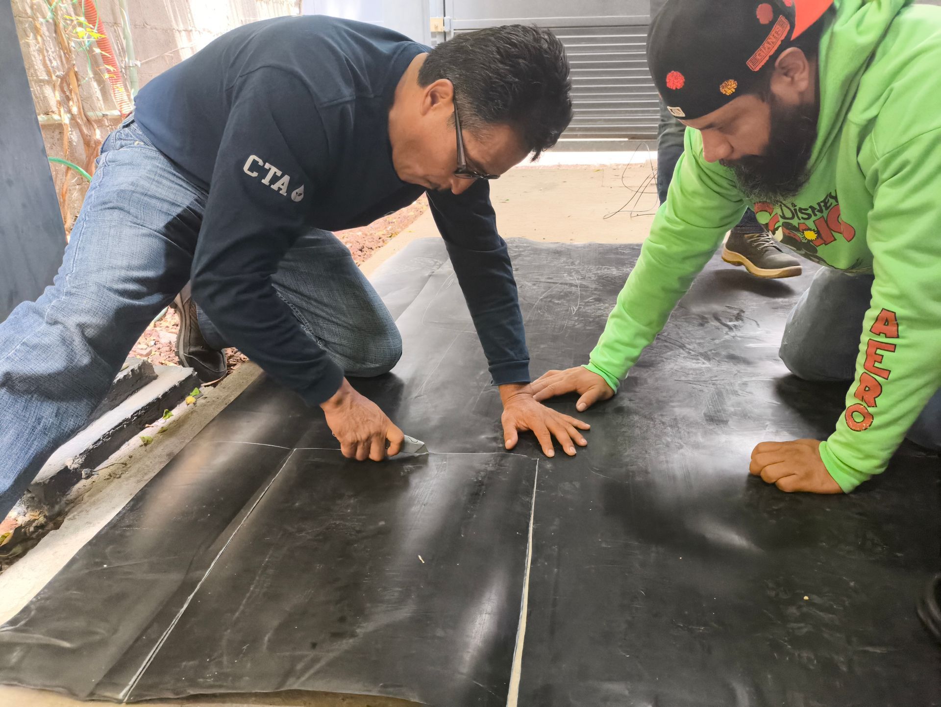 Dos trabajadores se arrodillan sobre un material de techo negro, alisando las juntas durante la instalación.