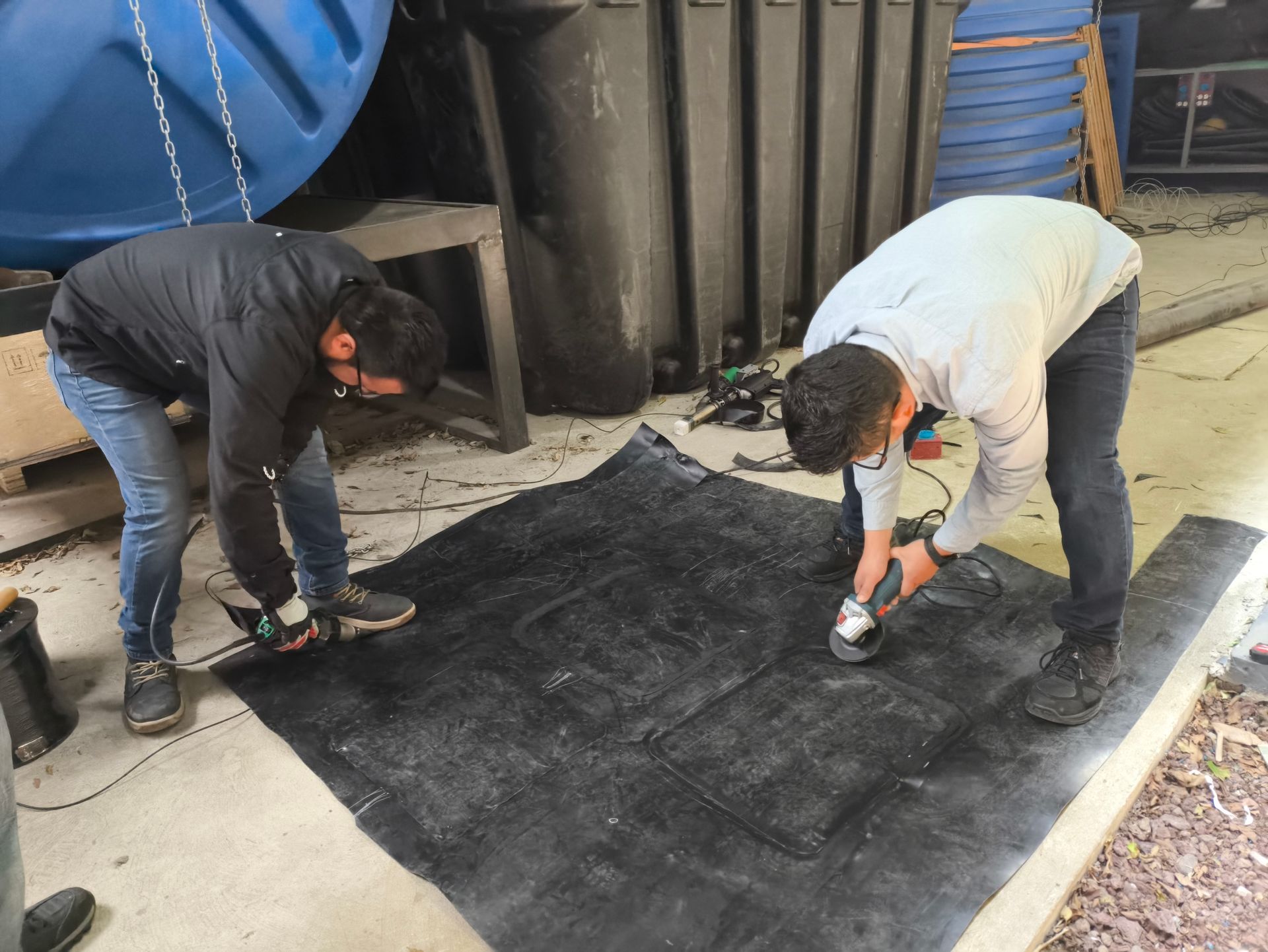 Dos trabajadores utilizan herramientas para reparar una gran alfombra negra al aire libre, cerca de tanques industriales.
