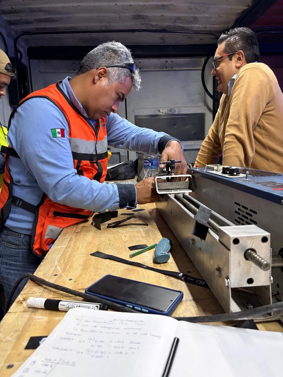 Dos trabajadores inspeccionan el equipo dentro de un camión; uno de ellos lleva un chaleco de seguridad naranja y utiliza herramientas.