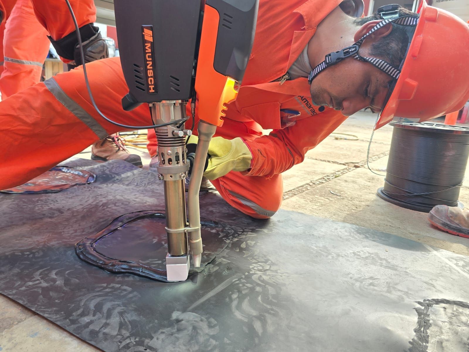 Trabajador con equipo de seguridad naranja utilizando una herramienta eléctrica para reparar pavimento o material de techado al aire libre.