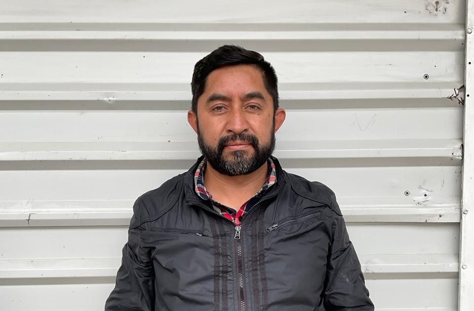 Hombre de pie contra una pared de chapa ondulada blanca, con una chaqueta oscura y mirando al frente.