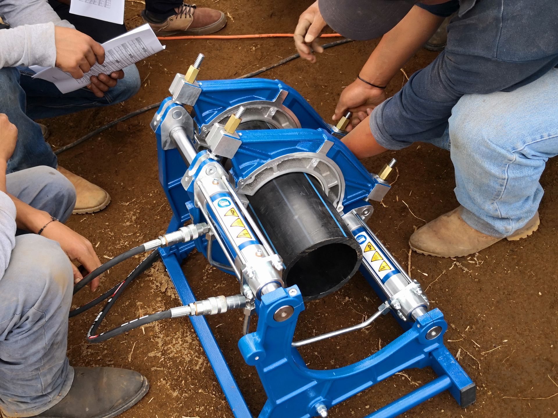 Trabajadores ensamblan una máquina de metal azul sobre un suelo de tierra, apretando con las manos las piezas alrededor de un cilindro negro.