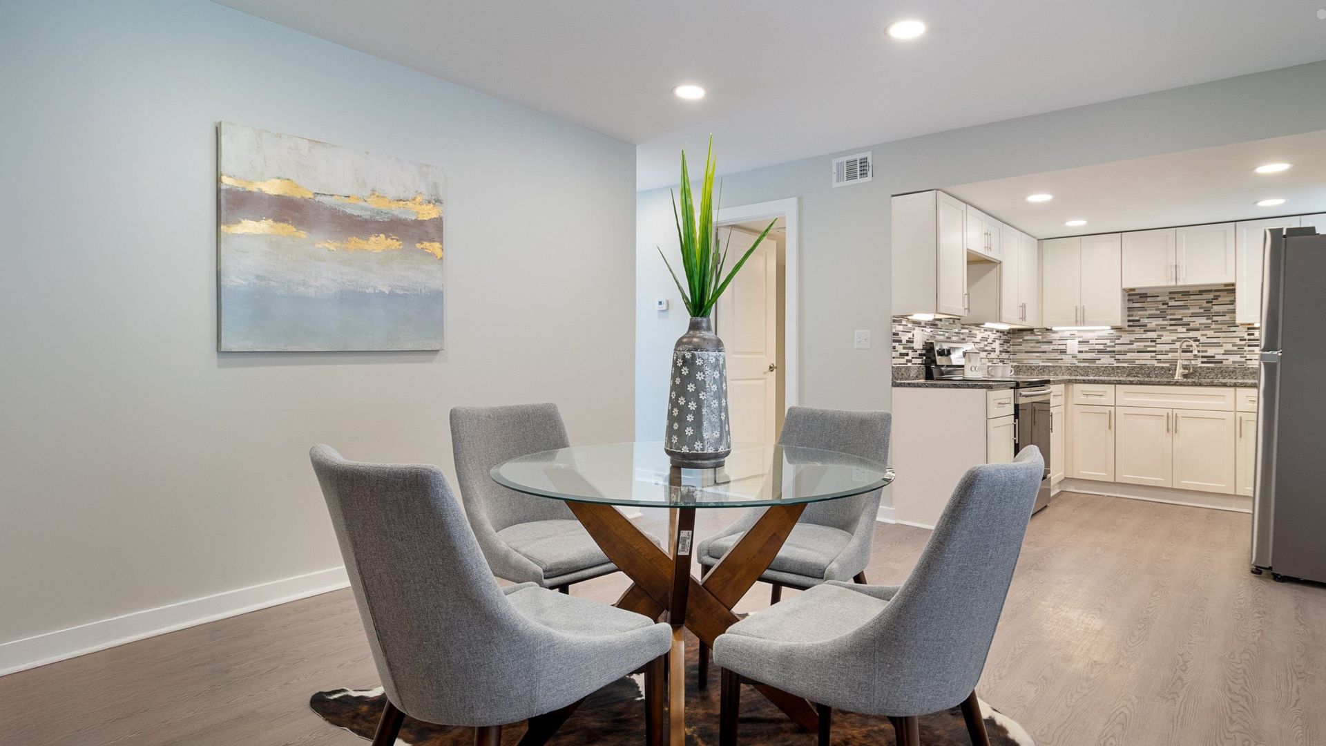 Modern dining nook with glass table and gray chairs, opening to a bright kitchen with white cabinets.