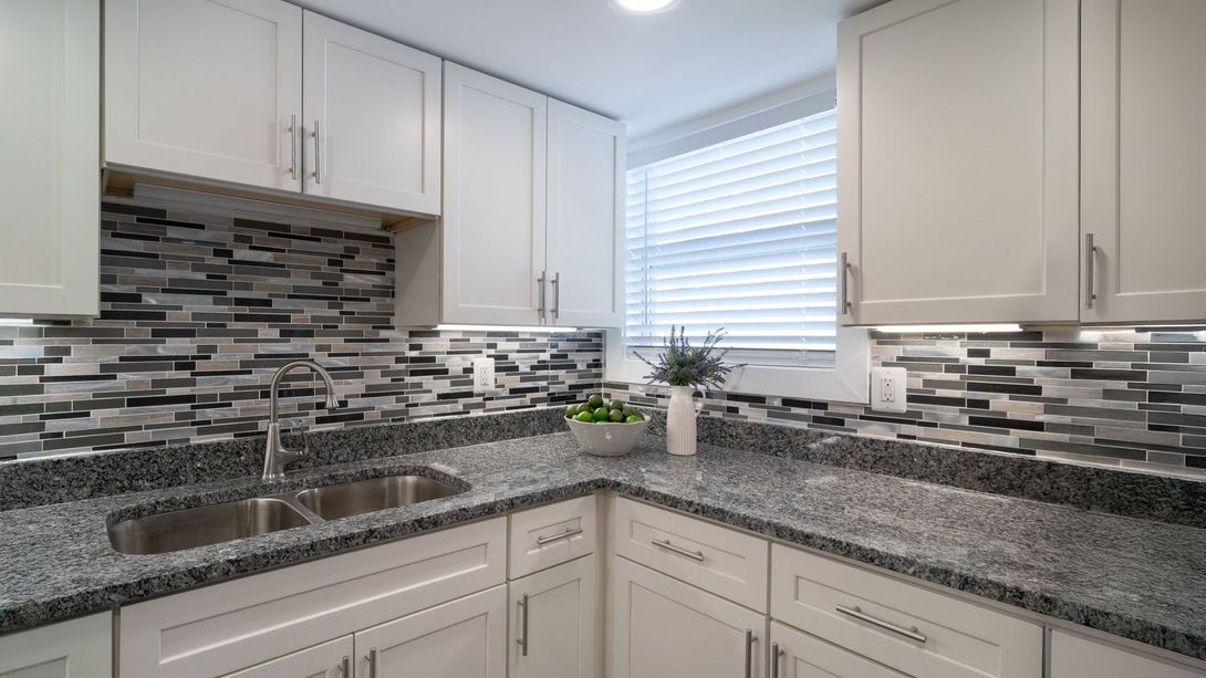 Bright modern kitchen with white cabinets, gray mosaic backsplash, granite counters, and a window over the sink
