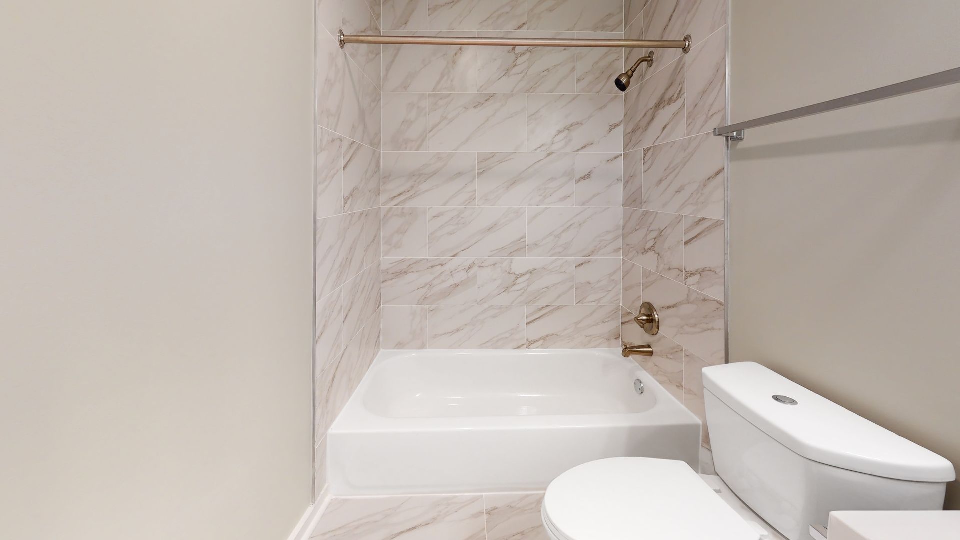 Small bathroom with white tub, tiled shower wall, gold shower rod, toilet, and towel bar.