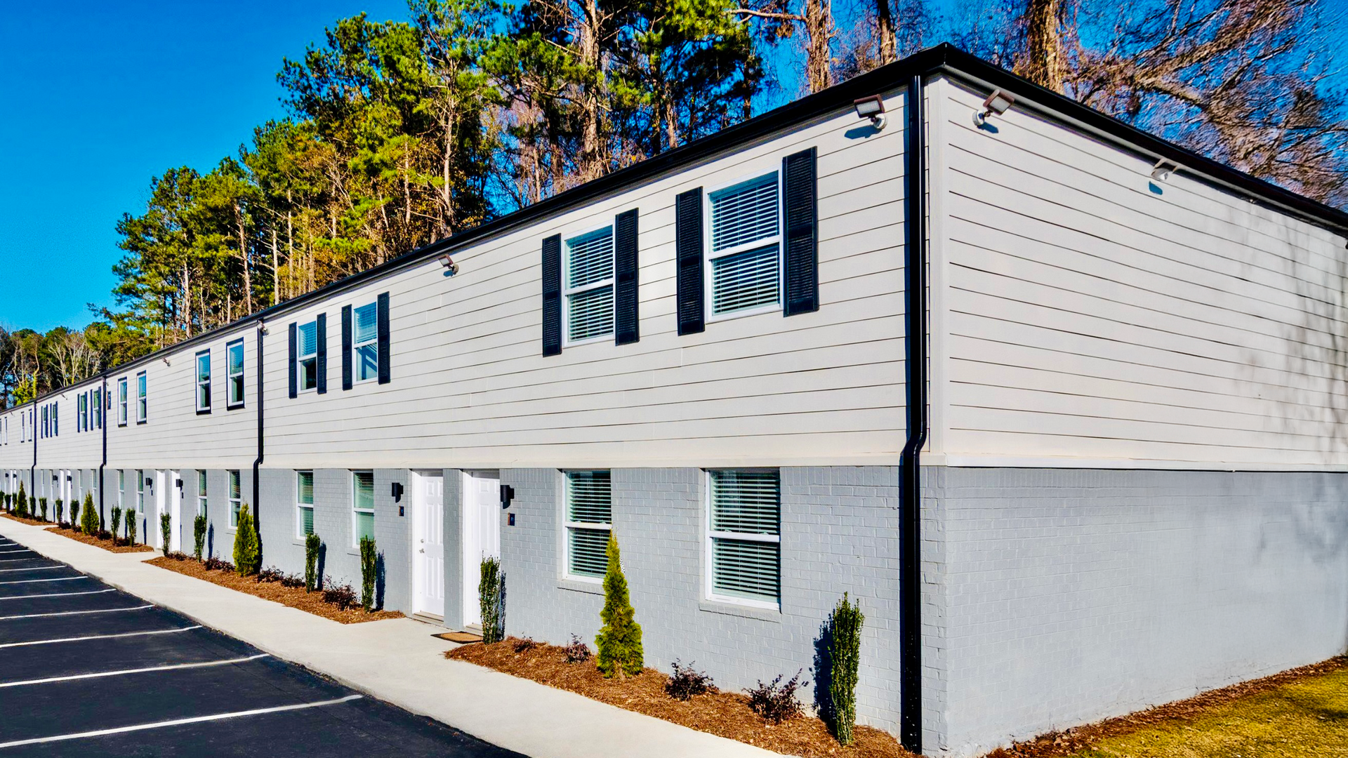 Two-story white apartment building with dark shutters along a paved street on a sunny day