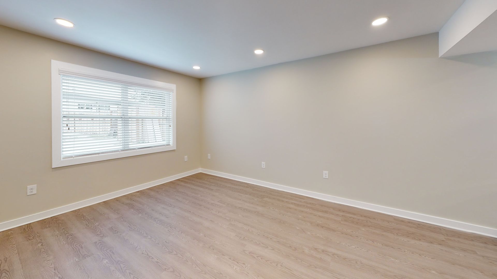 Empty beige living room with recessed lights, wood floor, and a window with blinds