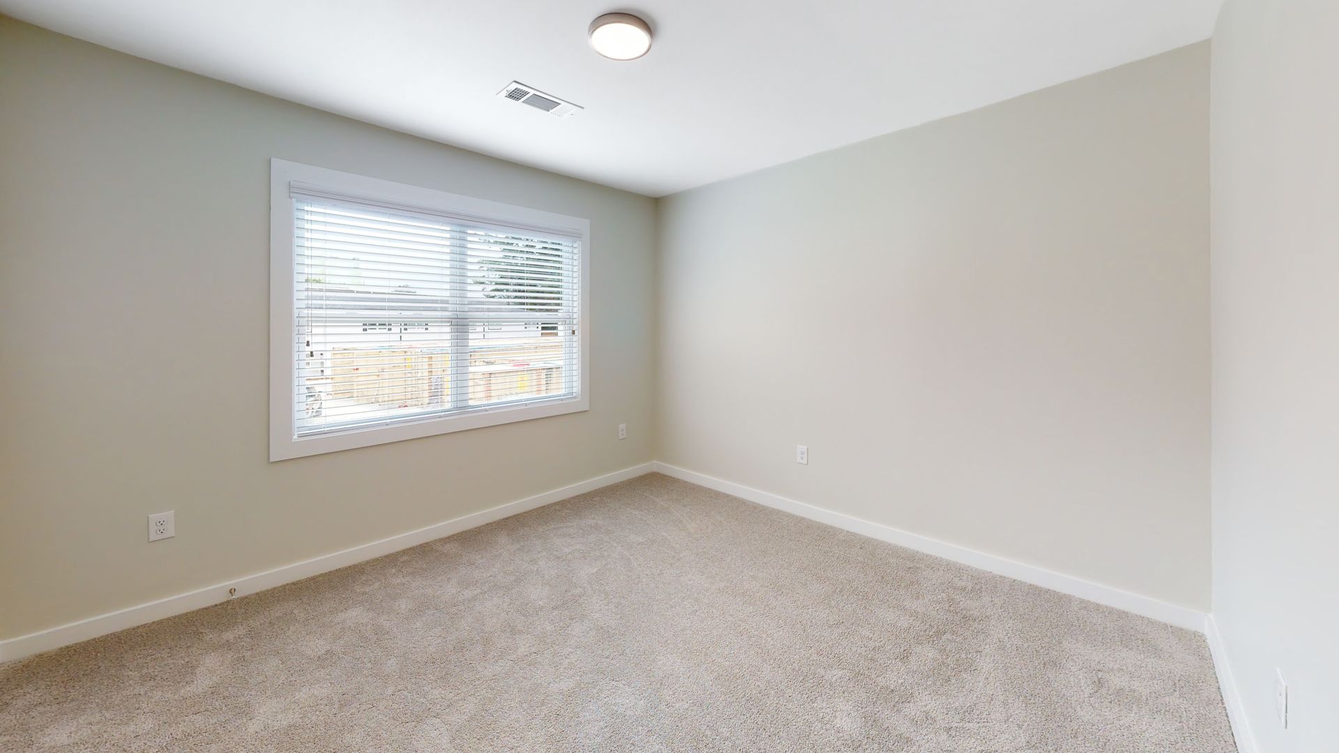 Empty beige bedroom with carpet, window blinds, and ceiling light.