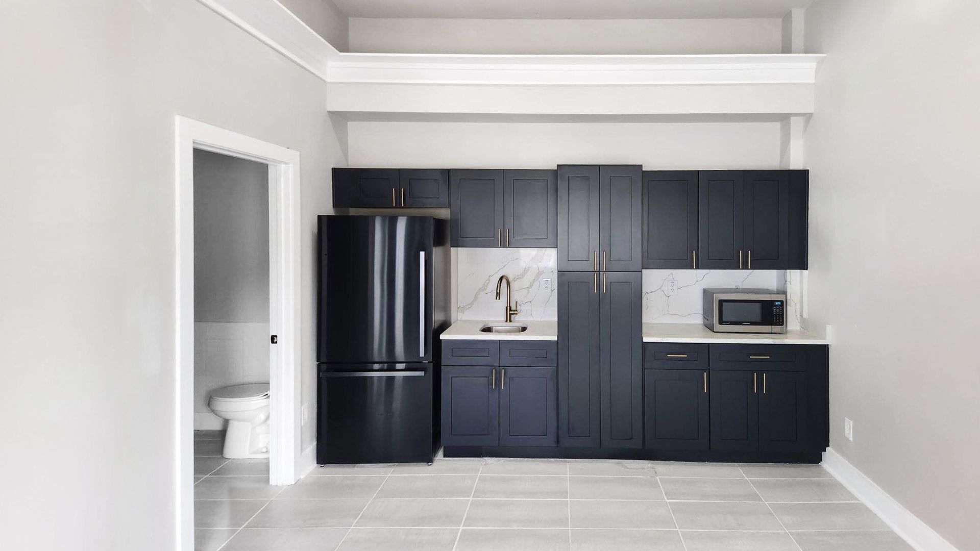 Modern kitchen with dark cabinets, black refrigerator, white countertop, and a bathroom visible through an open doorway.