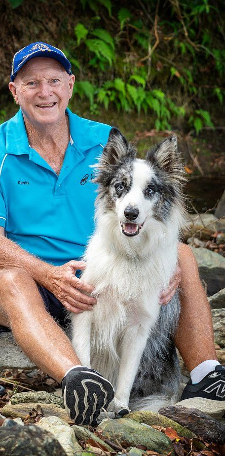 Man in blue shirt and baseball cap sits with a blue-merle Border Collie on rocks near a stream.— Canine Training School in Trinity Park, QLD