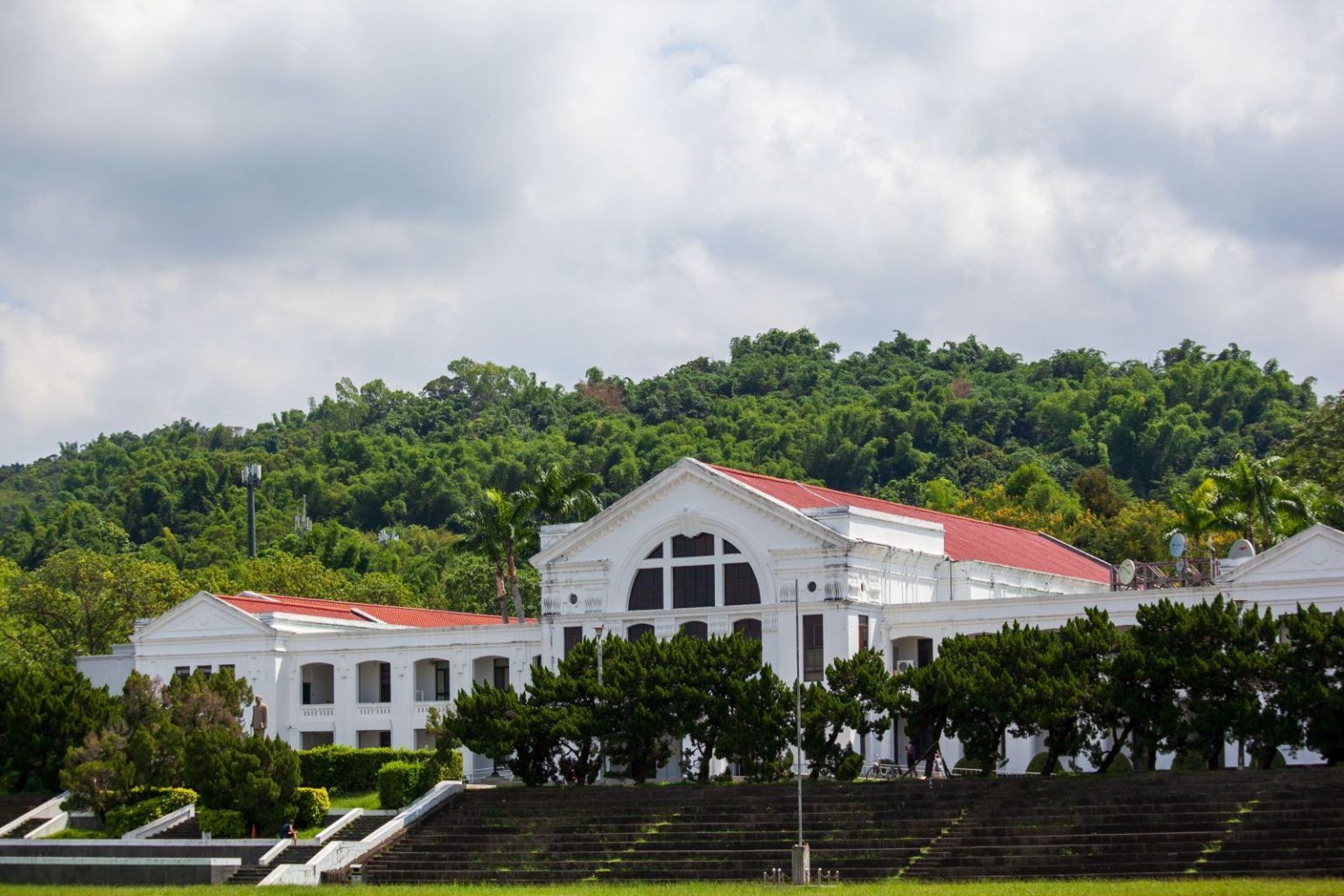 山坡上,一座白色殖民風格的建築,紅色的屋頂,背後是綠樹成蔭,天空陰雲密布。