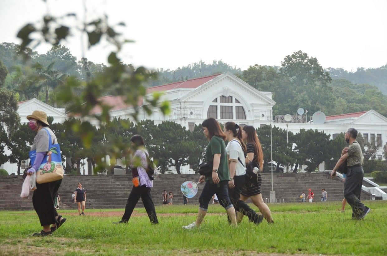 人們走過一片草地,草地前是一棟白色建築和樹木。