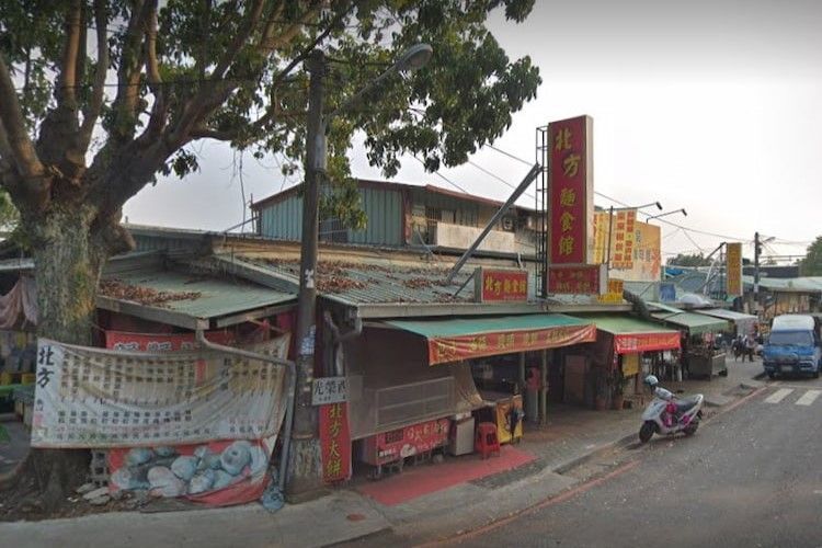 街邊一家中國餐館，紅色招牌，周圍綠樹成蔭，斜坡上停著摩托車。