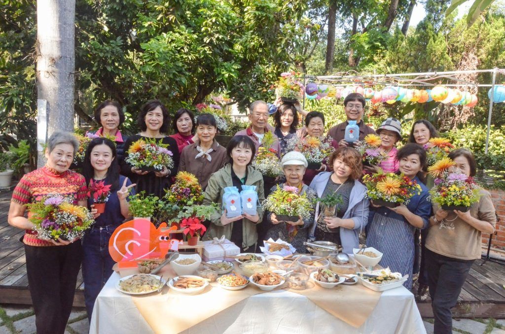一群人在戶外慶祝聚會上,手捧鮮花,圍坐在擺滿食物的桌子旁合照。