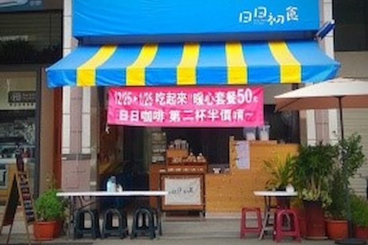 店面很小，有藍黃色的遮陽篷和粉紅色的中式橫幅，外面擺放著凳子、桌子和盆栽植物。