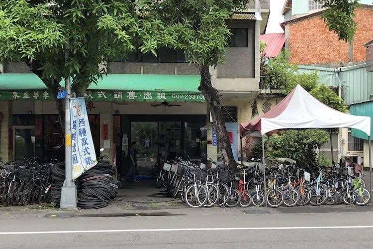 街邊自行車店,許多自行車停放在樹蔭下,還有一個白色遮陽篷。
