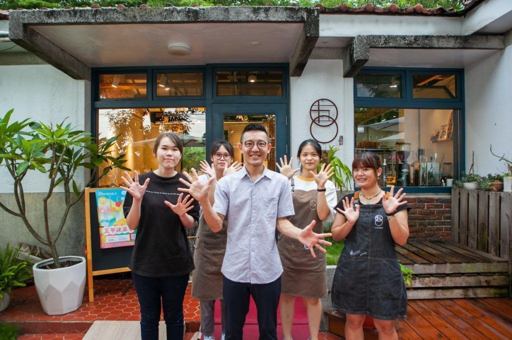 四個面帶笑容的人舉起雙手，在一家有著綠色遮陽棚的咖啡館外合影。