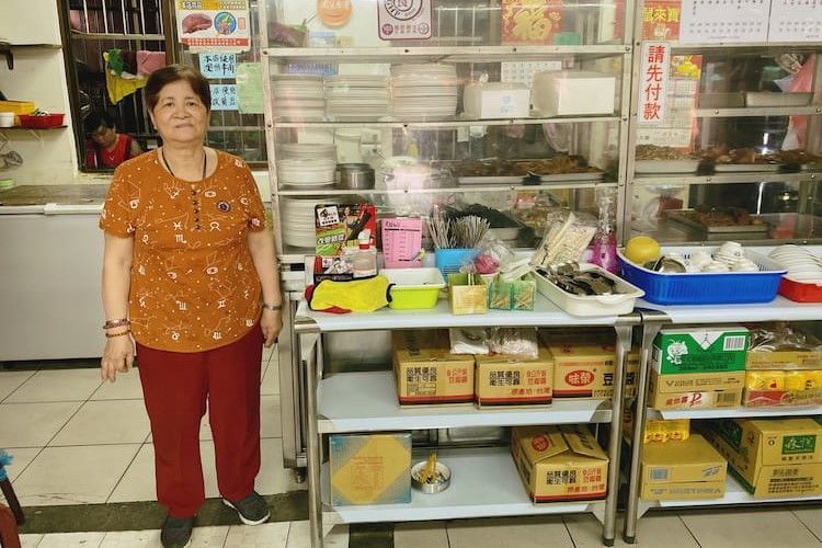 一位女士站在一個小型市場櫃檯旁，櫃檯上擺放著農產品、籃子和盒子。