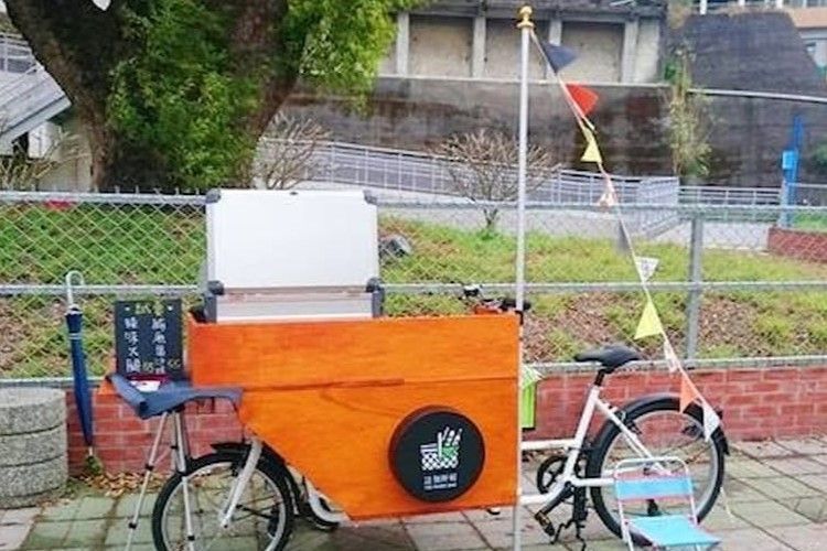 一輛木製貨運三輪車，車上插著一面小旗，停在戶外區域的柵欄旁。