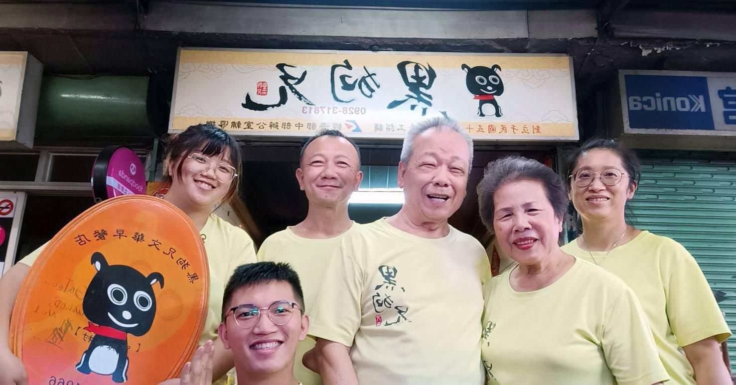 五個身穿黃色襯衫、面帶笑容的人在一家店面外擺姿勢拍照,店面上方有一個招牌。