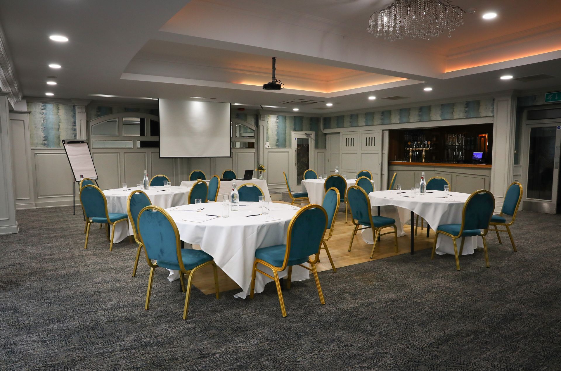 Empty banquet hall with round tables, white tablecloths, teal chairs, and a projector screen at the front