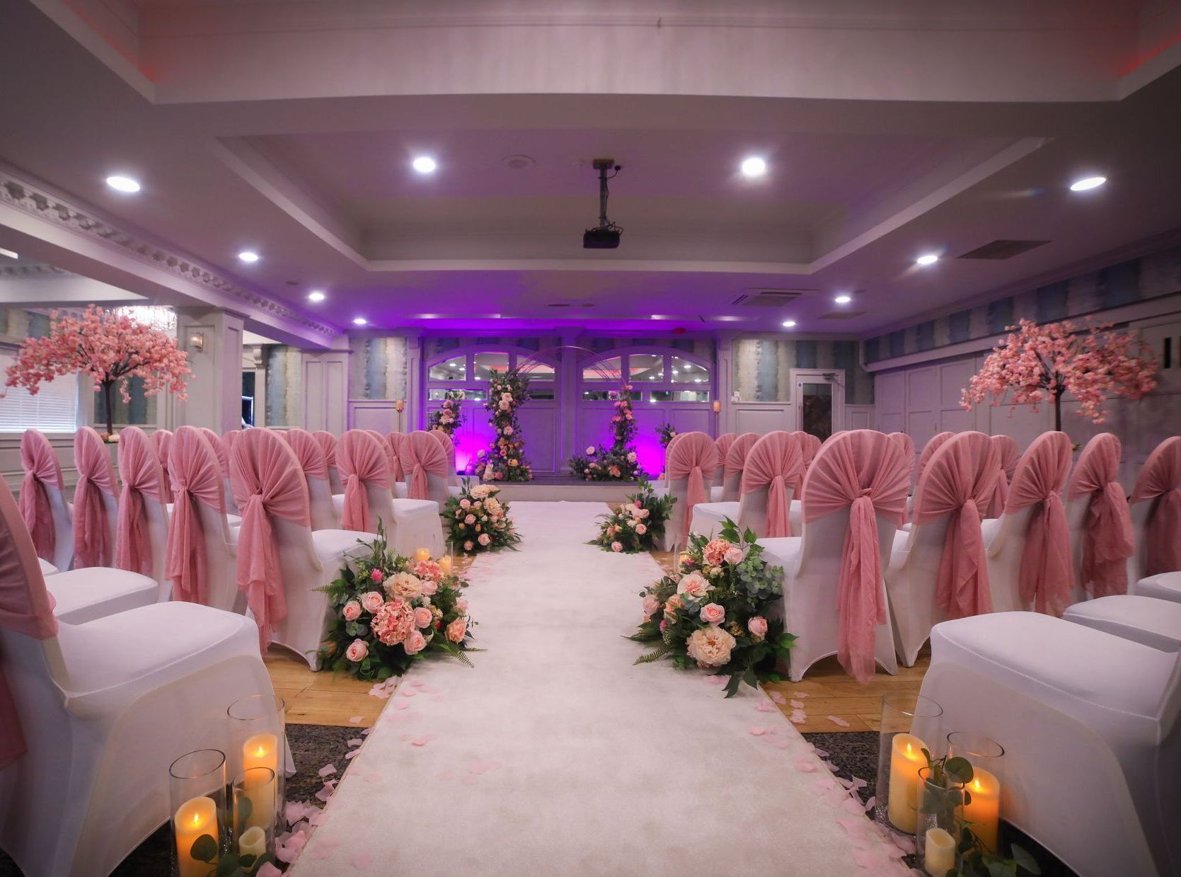 Decorated wedding aisle with pink chairs, floral arrangements, and purple uplighting in a reception hall