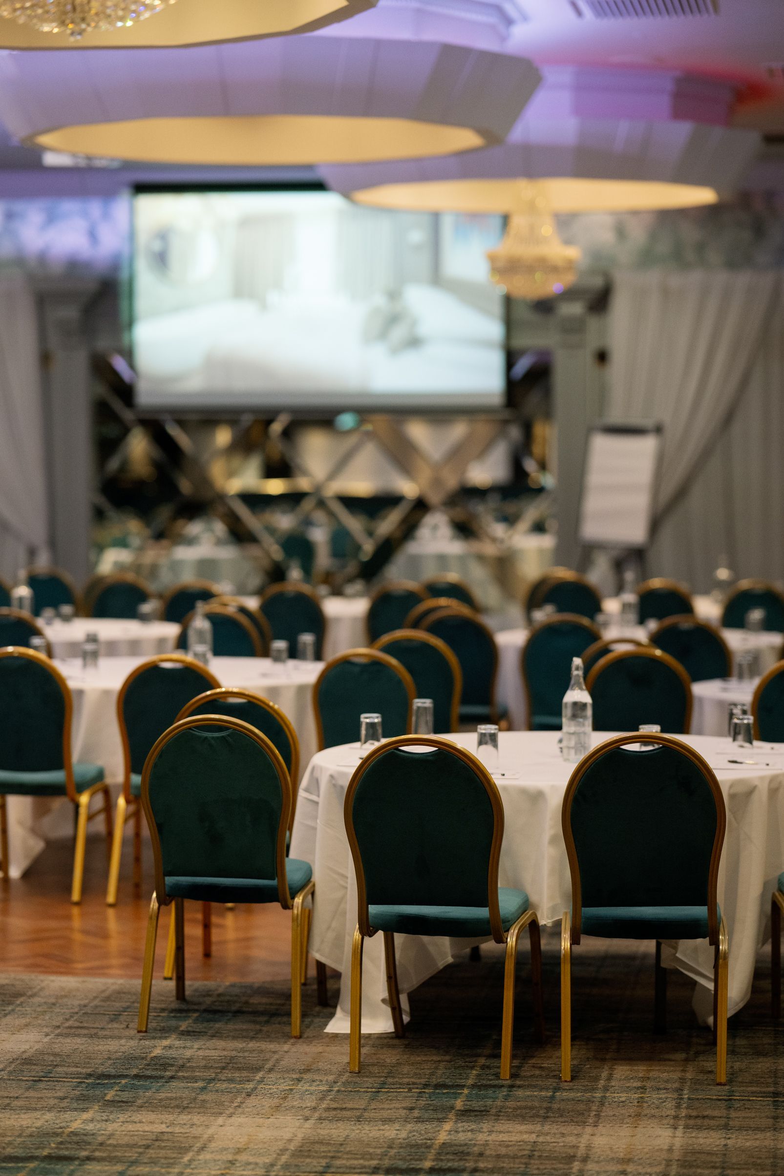 Elegant banquet hall with round tables, green chairs, and a large screen at the front for a presentation